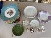Top view showing stacked colorful scalloped plates, white plates, small floral plates, green saucer, white-purple plate, and brooches on table