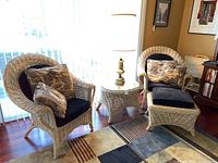 Two matching rattan chairs with black cushions, patterned throw pillows, a matching rattan ottoman with cushion, a glass-topped rattan side table, and a tall brass lamp.