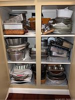 Three shelves with stacks of metal baking pans, glass and ceramic plates, bowls, and trays filling shelves tightly.