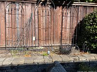 Group of metal garden trellises and plant cages arranged along a garden bed in front of a wooden fence, including tall rectangular trellises and cone shaped cages.