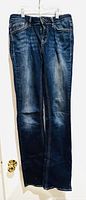 Front view of dark denim jeans hanging on hanger separated by door knobs. Shows worn faded detailing on thighs and knees, classic five-pocket style.
