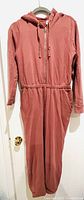Full view of dusty rose colored romper/jogger with hood, zipper front, and elastic waist hung on hanger.