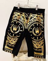 Black youth shorts with metallic gold graphic patterns and crown emblems seen from front on hanger