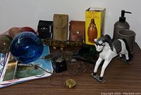Photo of assorted decorative and collectible items displayed on table, including leather cases, wooden horn, glass sphere, Aztec statue box, horse figurine, and other vintage items.