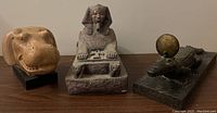 Three Egyptian-themed decor figurines on a wooden surface: left and center are hippopotamus heads and a seated pharaoh; right is a small crocodile on a base.