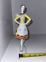 Porcelain figurine of a woman in traditional dress shown full height with measuring tape indicating scale; statue approximately 8.5 inches tall, standing on an oval base.
