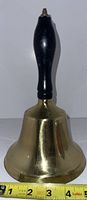 Full front view of large vintage brass bell with black wooden handle and a measuring tape showing approx 4 inches diameter