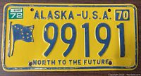 Front view of the yellow 1970 Alaska USA license plate numbered 99191 with stickers and state flag design.