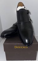 Pair of new men's Doucal's black leather double monk strap dress shoes displayed on brown Doucal's shoe box. Shoes stuffed with white paper to maintain shape, showing smooth polished finish and buckle details.