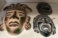 Three pottery tribal masks displayed on white shelf with different colors and tribal designs.