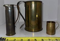 Set of three vintage trench art items: tall metal pitcher, large brass pitcher, and small brass mug on white background with measuring tape in front.