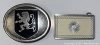 Front view of the two vintage belt buckles, one oval with a black lion on a shield, the other rectangular with a cream inset and sunburst pattern.