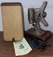 Vintage Keystone Model K-109 film projector with its original case, instruction manual and power cord displayed on wooden surface.