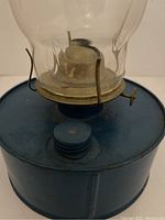 Side view of vintage lantern showing blue metal base, fuel container, and part of glass chimney with metal supports.