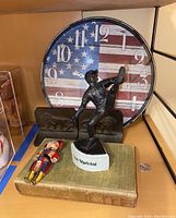 Wide shot of the metal bookends featuring a baseball player figurine, the American flag clock behind, old book under the doll