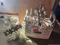 Two metal chandeliers placed on the floor next to a box, showing overall size, shape and chandelier design with candle bulb holders and crystal pendants.