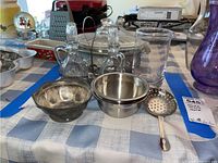 Two glass cruets with metal stoppers, metal bowl, two stainless steel bowls, glass pitcher, slotted spoon arranged on a checkered tablecloth