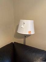 Full view of the floor lamp showing white tapered lampshade sitting on the nickel toned lamp stand in a corner.