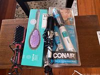 Three hair styling accessories on a wooden table: Conair Hot Air Curling Iron & Brush in plastic packaging, Dafni hair straightening brush in turquoise box, and a red/black Remington heated styling brush.