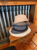 Four hats stacked on a wooden chair by a radiator: beige bucket hat, dark bucket hat with strap, black and white patterned fedora, white and beige straw fedora