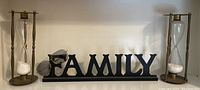 Two glass and metal hourglasses on either side of a black wooden FAMILY sign, also including aviator sunglasses on the sign.