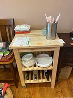 Overall view of the wood kitchen cart showing two shelves with items on the bottom shelf and top surface with cups and containers. One side of cart with wheels visible.