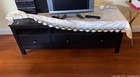 Front view of the black wooden TV console, showing three drawers with knobs and a lace cloth draped over the top surface. Scratches visible.