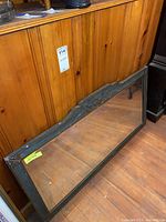 Vintage wooden framed mirror leaning against wooden panelled wall, showing wood grain floor reflection and frame details.