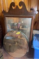 Two mirrors leaning against a wooden paneled wall: one rectangular with a dark wooden frame featuring decorative top, and one round glass mirror.