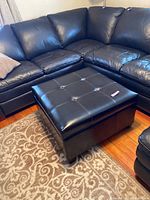 Black leather-like square ottoman with tufted top and buttons, placed on patterned carpet, next to sectional sofa.