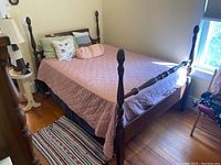 Side view of the full size wooden bedframe with pineapple finials, mattress covered with light pink quilted bedspread, and pillows in a bedroom.