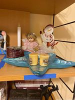 Shelf display showing metal snowman figure in white with red accents, angel doll in pink dress, two votive candles on blue star dish, and roll of burgundy ribbon.