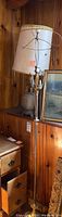 Floor lamp with metal frame and no shade, positioned next to table lamp and painting against wood paneling