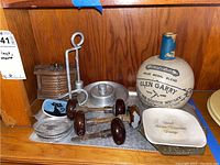 Metal tray, vintage wooden and cork coasters in curved wooden case with brass details, metal candle holder with snuffer, 6 metal coasters, ceramic whisky flask, ceramic dishes, and vintage door knobs on wooden shelf.