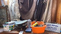 Overview of lot items on wood surface against fabric and wood background, including first aid kit box, orange wash bin with tools, brass mortar and pestle, metal light shades, tape measure, and Philips light bulbs.