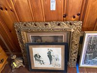 Two framed vintage pictures leaning against a wooden paneled wall. One ornate gold frame with vintage bank advertisement and a simpler black frame with an illustration of three people.