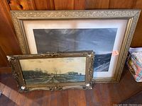 Two framed paintings leaning against a wooden wall on a wooden floor. Larger frame contains ocean scene with sailboat, smaller contains pastoral landscape. Both frames are ornately carved and show damage.