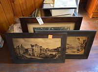 Three vintage framed prints stacked leaning against a wooden wall, showing dark wooden frames and visible wear, tear, and staining on pictures, some cracked glass evident