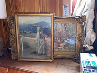 Two framed paintings leaning against a wooden panel. The front painting depicts a girl standing by a statue on a terrace with mountains behind. The back painting shows a nature scene. Both frames are ornate and chipped.