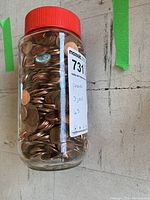 Side view of clear plastic jar filled with many pennies and red lid, showing label with lot number and penny count notes.