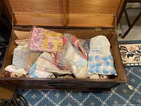 Wide view of wooden chest filled with assorted fabric remnants, yarn, thread, and table linens mostly in plastic bags.