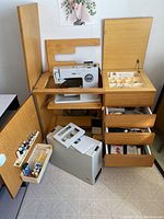 Full view of sewing machine integrated into wood sewing cabinet with open drawers and doors showing sewing notions and accessories.
