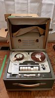 Photo showing vintage reel to reel tape recorder inside its carrying case with two reels loaded and control buttons on front panel.