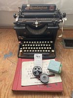 Full front view of Remington 12 typewriter showing keys, brand label, included ribbon, book, and instruction sheet in front.