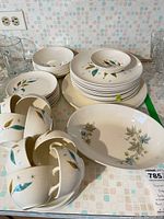 Photo of stacked plates, bowls, coffee cups, and saucers with green and gold leaf and starburst design on cream background.