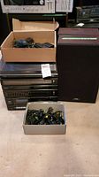 Full stereo system with turntable on top, cassette deck, radio tuner, equalizer, and control buttons. Two large speakers to the sides. Cardboard boxes on top and in front holding cables and accessories.
