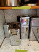 Five metal canisters placed on tiled kitchen counter. Four have labels for sugar, coffee, flour, and cocoa. One unlabeled canister is behind the sugar canister.