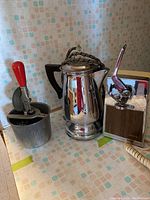 Photo shows three kitchen items together: manual can opener with red handle, shiny metal coffee percolator, and a hand-powered food mixer with a red crank and woodgrain panel.