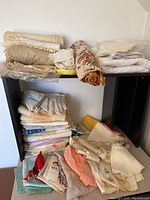 Wide view of vintage linens piled on a dark wooden furniture piece including folded towels, tablecloths, doilies, handkerchiefs, and placemats in beige, cream, and some colored fabrics.
