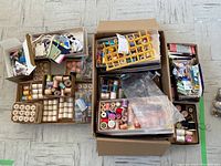 Overview photo showing multiple cardboard boxes and trays filled with vintage sewing notions including spools of thread, buttons, lace, and beads arranged on a tiled floor.